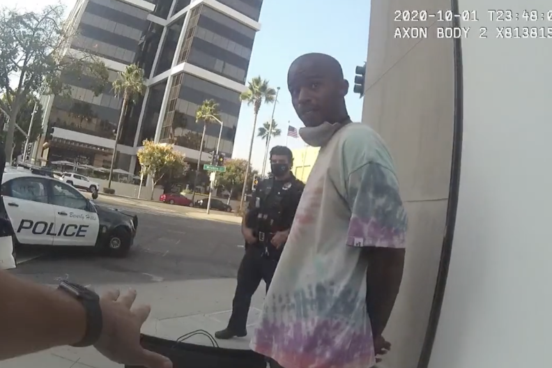 A Black man with a mask around his chin and his hands behind his back speaks to a police officer with another officer in the background.