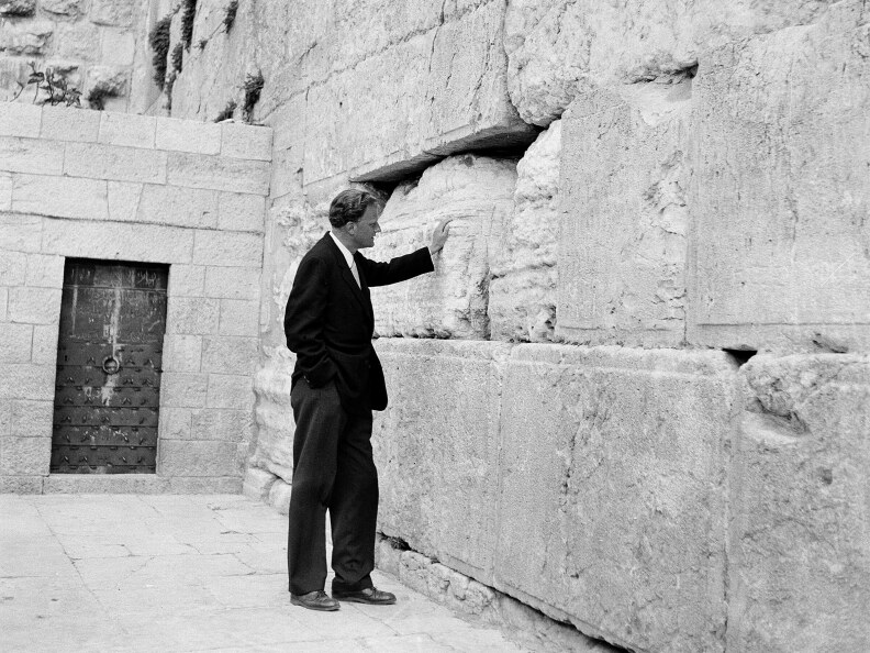 A black and white picture shows a tall white man in a dark suit with his left palm flat against an ancient stone wall.