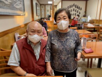 A portrait of David and Kyung Han. The couple are posing inside their Olympic Restaurant. A server is working at a table in the background.