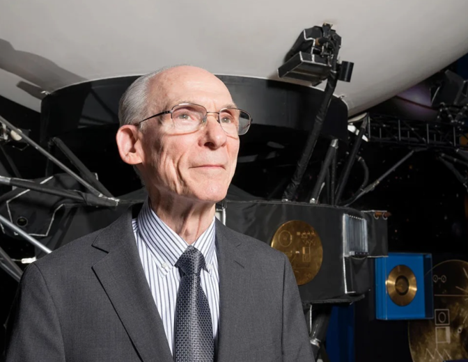 An older adult man stands in front a space craft wearing a great suit change, striped shirt, and grey tie. The man has glasses and is looking past the camera. 