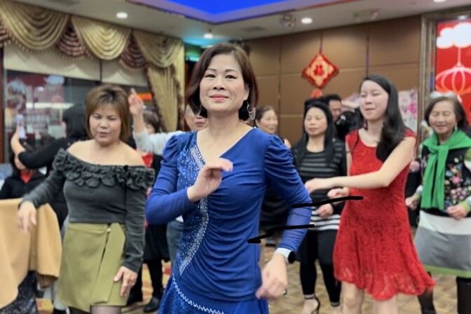 A group of a half dozen Asian American women do a line dance in a Chinese restaurant. In the foreground is a woman with a reddish brown bob and blue sparkly dress.