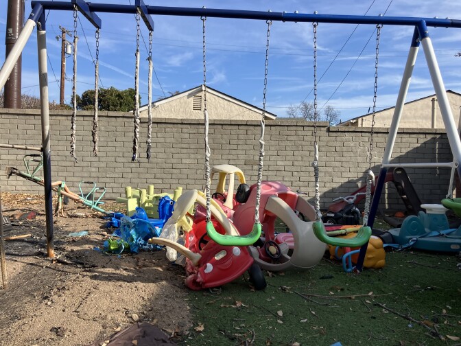 A blue and white swing set with green swings. Half the ground on the left side is covered in sand. The right side is covered in green fake grass. There are three swings on the swing set, but only the middle and right hand one are in tact. The swing on the left has just chains and no swing seat. The chains look charred. Behind the swing set, a children's red plastic truck is semi-melted. A tangle of other plastic colorful toys are behind it. Branches and ash is strewn across the ground.