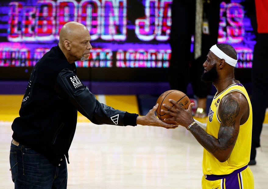Two Black men on a basketball court, one passing the ball to another. The man on the left appears to be in his 50s and is dressed in civilian clothes. The man on the right appears to be in his 20s or 30s and is dressed in a yellow uniform with a white sweatband on his head.
