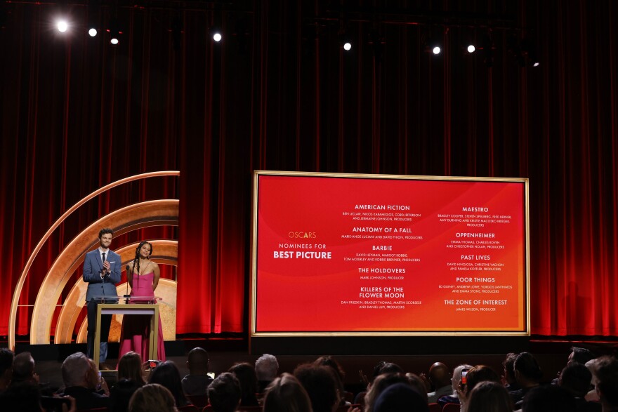 Two people stand at a lectern to the right of a big board listing 10 films nominated for best picture.
