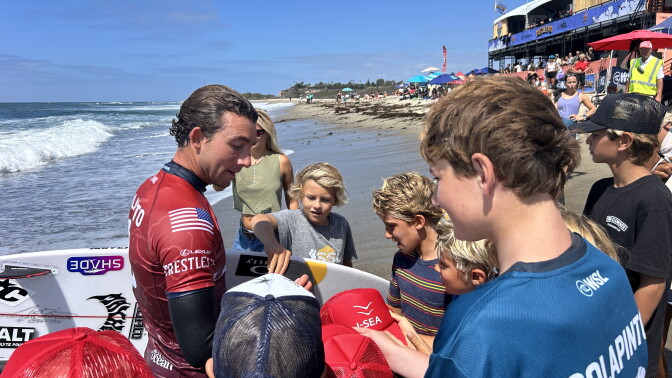 A wetsuit-clad surfer signs hats and a surfboard for a group of children gathered on the beach near a competition venue. One of the kids has a shirt that reads "Colapinto" on the back. 
