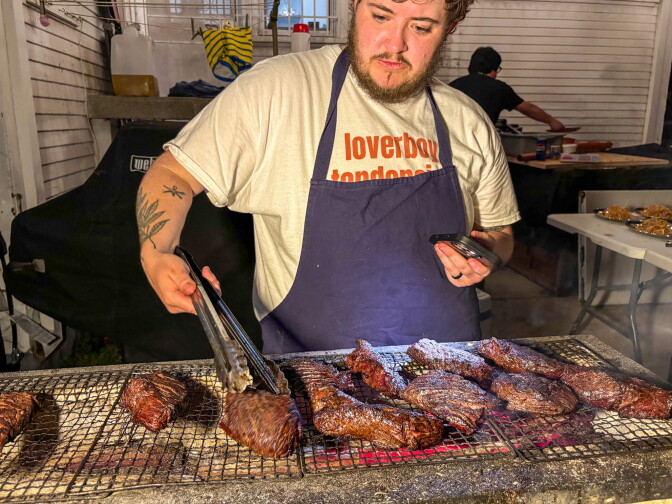 A man with a light skin tone and beard, wearing a navy apron over a white t-shirt, uses tongs to tend to a full grill of hanger steaks over glowing charcoal in the backyard of a Long Beach home at night.