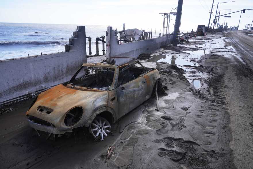 A small two-door car sits gutted and surrounded by mud and debris near a coastline.