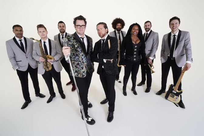 Nine musicians stand against a white backdrop posing with instruments and microphones. 