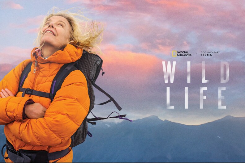 A blond woman in a orange jacket wearing a black backpack, mountains and a colorful sky in the background. Wild Life film title on the right.