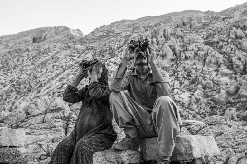 Two people sit on rocks using binoculars to look up