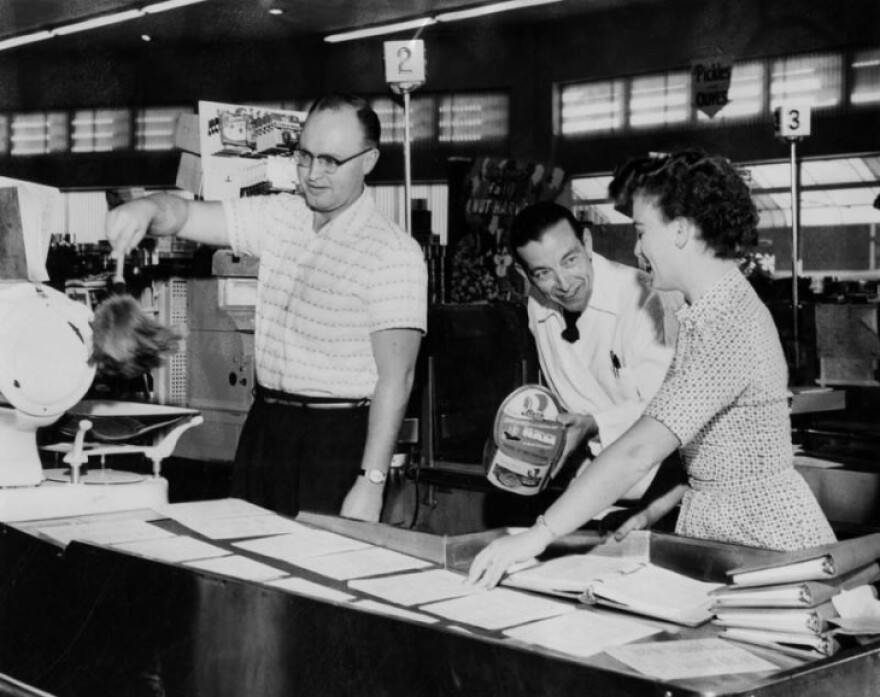 A black and white photo has people at a counter with one man dusting with a feather duster.