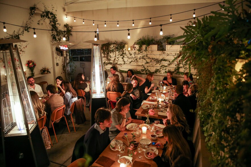 An overhead view of a busy restaurant's patio at night; there are tables filled with people laughing and chatting, with candles on each table and greenery all around