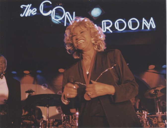 An older woman with light brown skin and blond curls to her shoulders stands on stage smiling with a microphone in front of drum cymbals and a lit up sign that appears to spell out "Conga Room" 