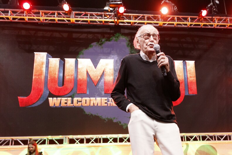 An older man, Stan Lee, holds a microphone on stage in front of a screen promoting the film "Jumanji: Welcome to the Jungle." Another person is seen in the background on the lower left.