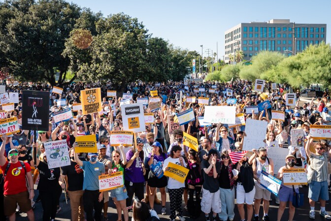 A crowd of people gathers, holding yellow signs that read "Stand With Animation!" and handmade signs. The ground stretches into the background, where trees that frame the street. One building is visible in the back right corner of the frame.