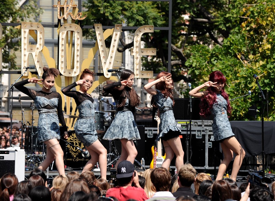 A group of five Asian women in blue dresses are lined up on stage striking a pose, performing in front of a daytime crowd. Behind them are the letters for the Grove shopping center.