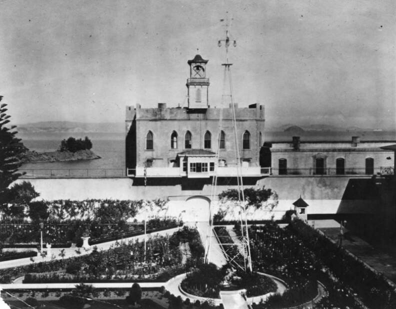 a building with a tower in the background. in the foreground are gardens, walkways and a tall telephone tower.