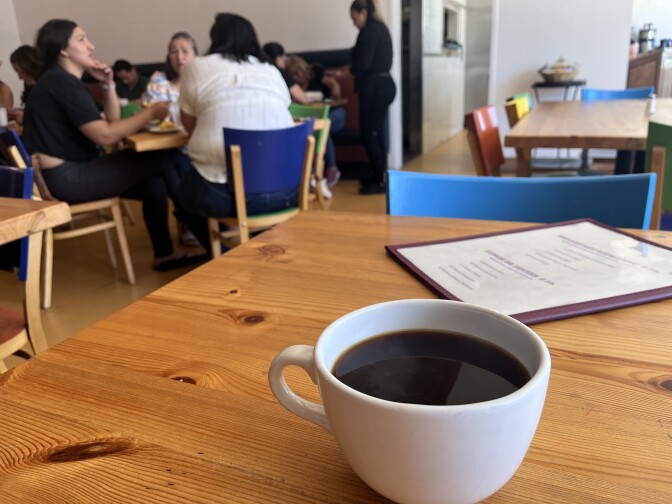 A mug of dark coffee sits on a table with a menu behind it. There are patrons sitting at tables in the background.