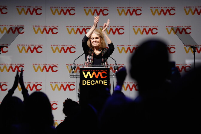 A woman stands at a podium, raising her hands above her head, clapping. In the foreground a crowd of people is people pictured in silhouette.
