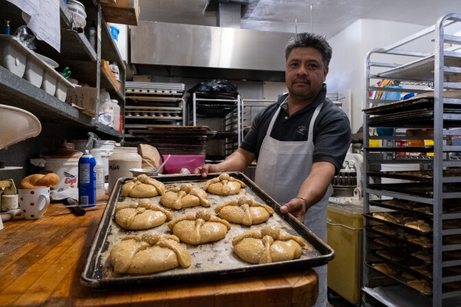 A man with medium-dark skin and black hair, streaked with gray, is wearing a black short-sleeved polo shirt and a white apron inside a stainless steel bakery. He is holding a large metal baking tray filled with unbaked bun pastries, featuring an 'X' design on top and sprinkled with tiny white sesame seeds. 