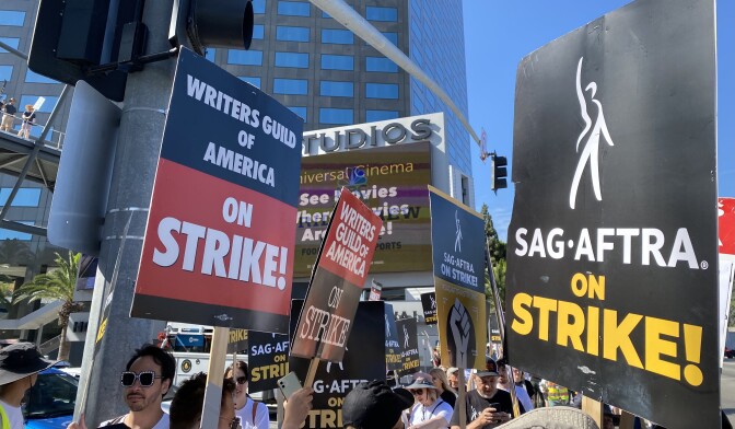 A sidewalk is thick with people carrying protest signs that say: "Writers Guild of America on Strike!" and "SAG-AFTRA on Strike!"