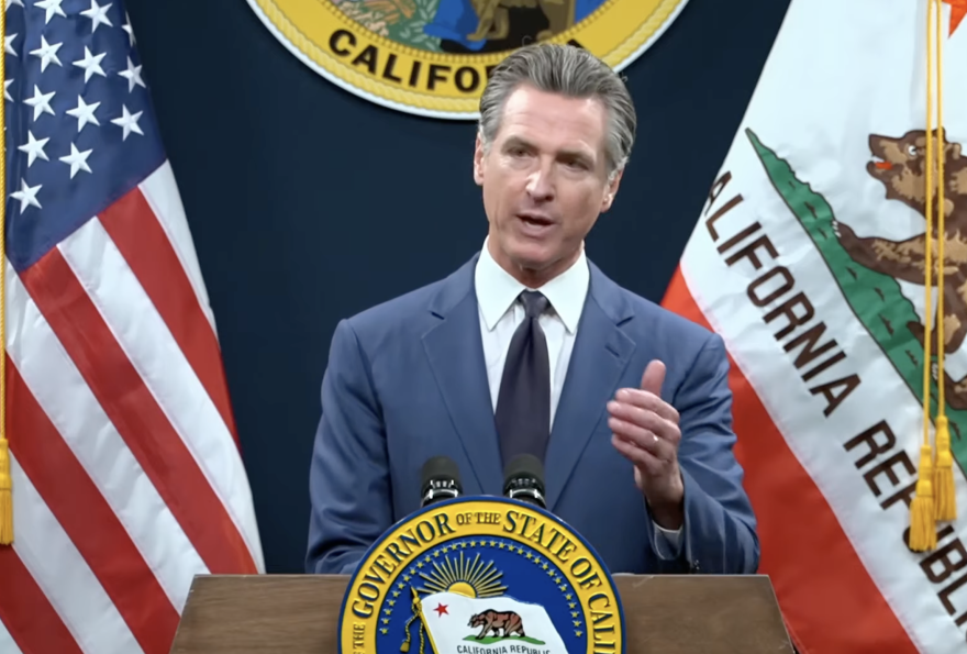 A man in a blue suit stands at a lectern with the seal of the Governor of the State of California.