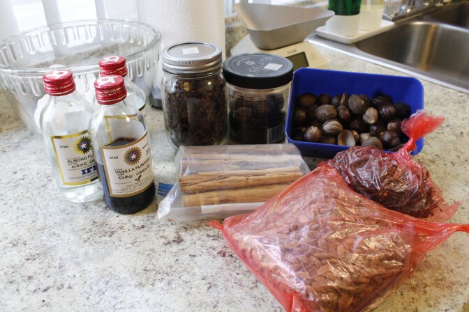 Three glass bottles with red caps are on the left side of the picture. On the right side two jars contain whole spices. A blue open tupperware contains nutmeg, while three plastic bags contain cardamom, cloves and cinnamon.