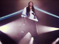 Rihanna stands center stage bathed in light wearing black loose pants and a white top.