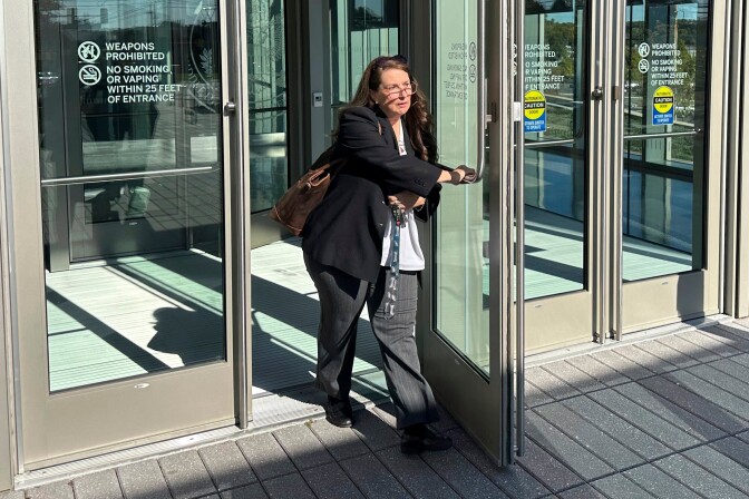 A woman wearing grey pants, a black jacket and carrying a brown bag pushes the glass door open of a building.