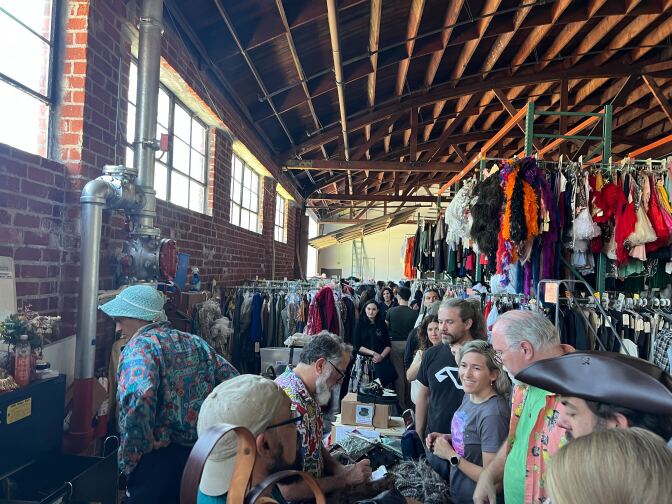 A picture of at least eight people, mostly with lighter skin tones, stand in a line to buy things in a warehouse with racks of colorful skirts and petticoats and other articles of clothing hanging behind them. 