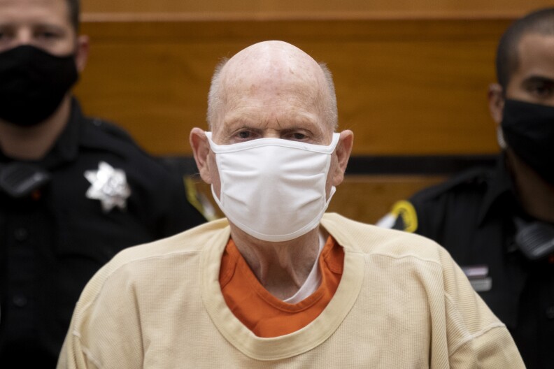 A elderly man with light tone skin and very little hair wears a white surgical mask in a close-up shot in a courtroom with uniformed officers behind him.