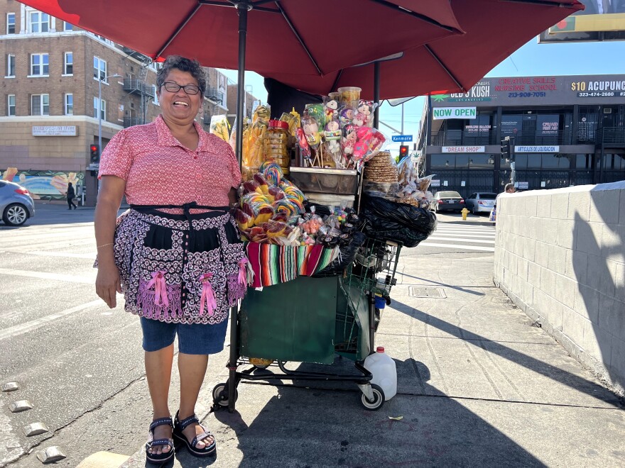 A Brown woman with short curly hair wearing a pink and black apron stand in front of a cart with an umbrella. On the cart, there are lollipops, an assortment of candies and chopped mangoes in plastic containers.