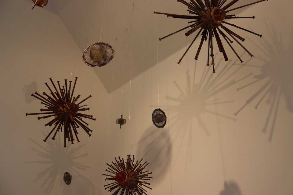 Several small sculptural objects are hung from a ceiling. They are spherical. Some have many stick-like pointers coming out from them, and others look like circles with smaller circles inside. 