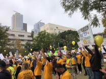 A crowd of people in yellow shirts with signs and yellow balloons cheer with hands up in a grass field in Grand Park in downtown L.A. as buildings loom in the background. 
