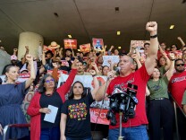 A group of people with varying skin tones raise their fists in the air. Many of them wear red shirts. Several people hold signs that say "educación, no deportación."