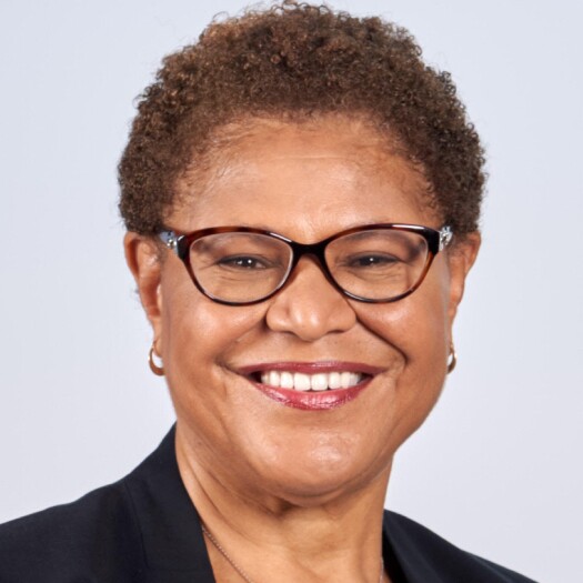 Headshot of Karen Bass. She has light brown skin and short, curly light brown hair. She is smiling and wearing a dark suit jacket or blouse, black and brown horn-rimmed glasses, and small hoop earrings.