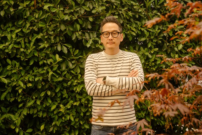 Glenn Kaino stands, arms folded, in a white and navy striped shirt, with a plant with green leaves behind him, one with red leaves in the foreground.