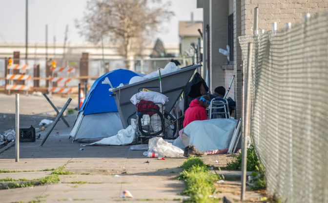 Trash is strewn around an overflowing dumpster on a sidewalk. A blue tent is beside the dumpster.