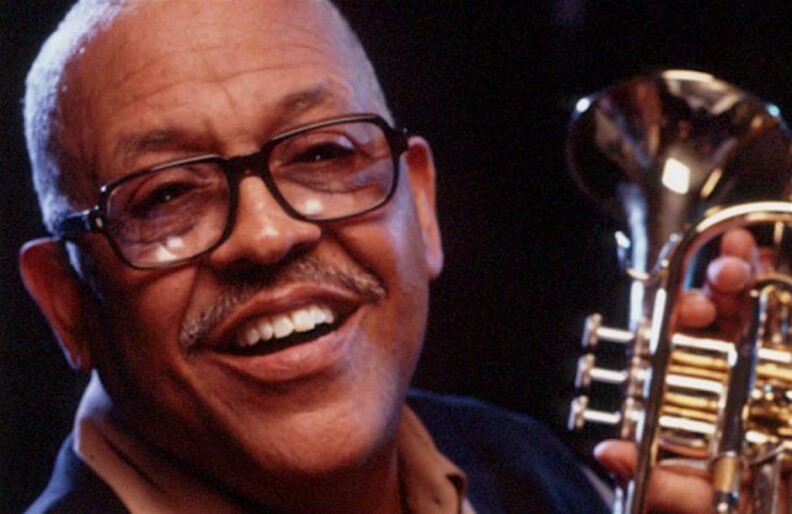 Bobby Bradford, a Black man wearing black-rimmed glasses, looks to camera while holding a trumpet.