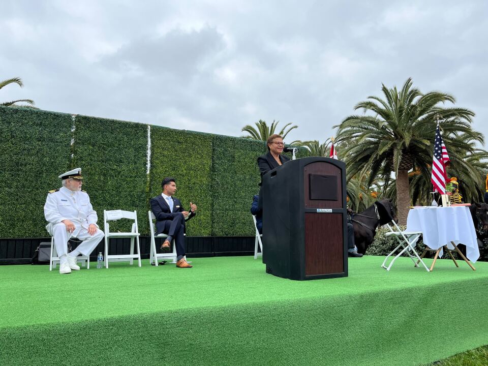On a stage covered in light green, a woman with brown skin tone wearing glasses stands at a podium. To the right of the image is a table covered in white. To the left are two men sitting with a white chair in between them. One of the men appears older, with light skin and dressed in an all-white uniform.