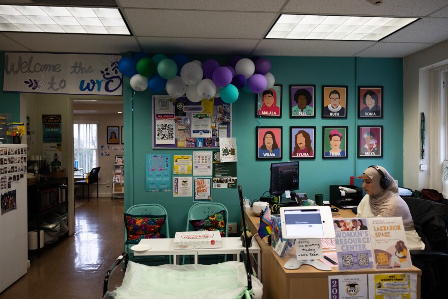 A feminine presenting person wearing a beige hijab sits a desk with various signs, a computer, and a iPad. The wall behind them is turquoise with art of various famous women. There's a door way with a sign that reads "Welcome to the WR"