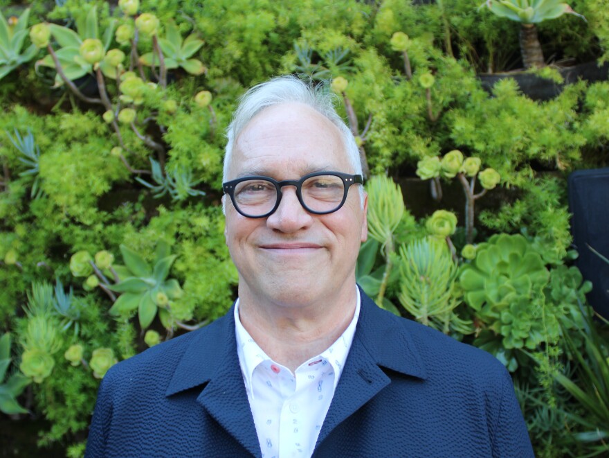 A man with white hair and glasses stands in front of greenery. 