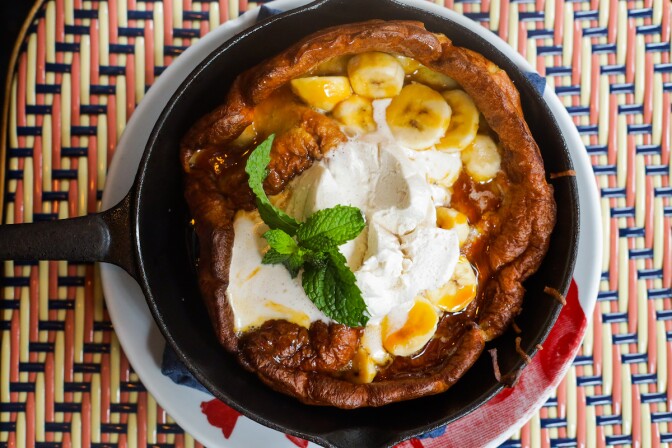 a skillet of a Dutch baby pancake with topped with bananas, whipped cream and a mint leaf.
