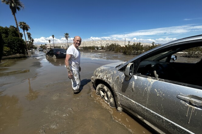 A middle aged, balding, light skinned man wearing a white t-shirt and mud-splatted white pants stands knee deep in mud beside a car stuck in the mud