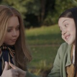 Amie Donald and Violet McGraw sit together outside with green grass and trees behind them. Donald sits on the left wearing a tan tailored dress with a large bow tie with blue, red, and gold stripes. The tails of the bow tie are long, falling down her shirt front. Her long blonde hair falls loose at her shoulders. McGraw wears a light green cardigan with tan overalls peaking through over a white t-shirt. Her long brown hair creates a straight curtain across the front of her left side. The two girls hold hands, with each of their thumbs raised, seeming to be playing a game of thumb war. Donald stares blankly past McGraw, her appearance robotic and artificial. McGraw focuses intently on their interlocked hands, her eyes are wide and a toothful smile across her face.