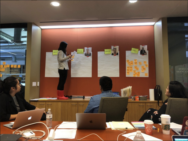 A woman stands on a counter and looks at four large pieces of paper taped to a wall while 3 people sit in chairs and listen to her