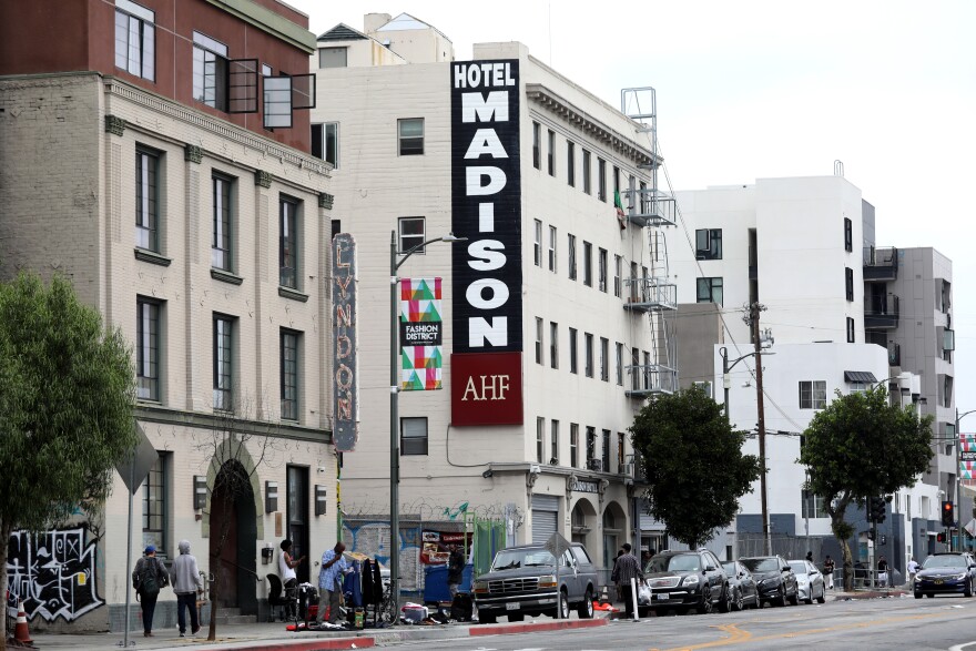A five-story building has a large painted sign reading: Hotel Madison AHF. People walk past on a street with parked cars.