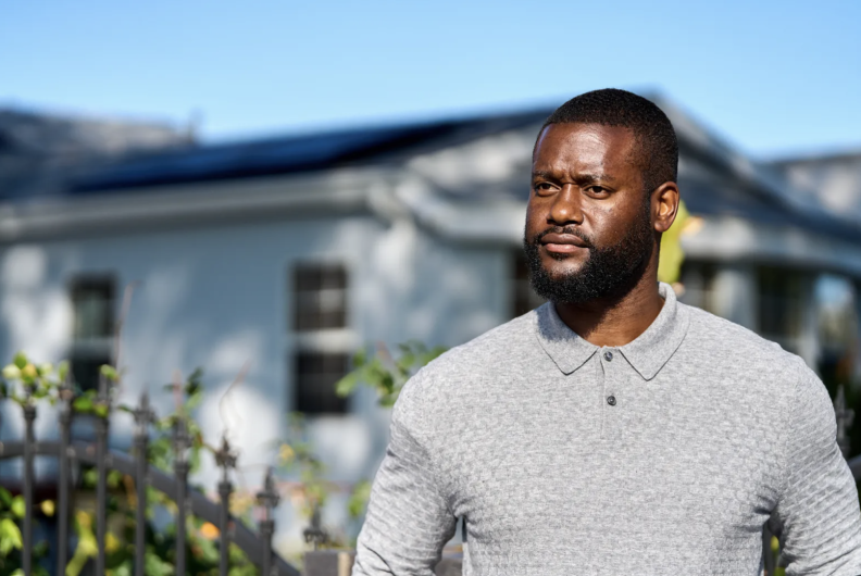 A Black man is standing for a picture during the day. He is looking to the left. Behind him is a house, out of focus.