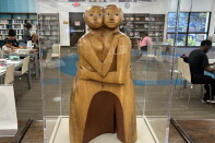 A wood sculpture of two people, they embrace and look at the viewer.