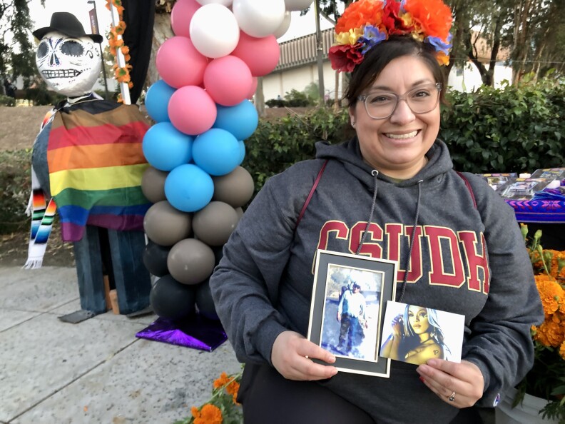 A light-skinned woman holds two photos of deceased people. She's in front of a Day of the Dead altar with gray, blue, pink, and white balloons.